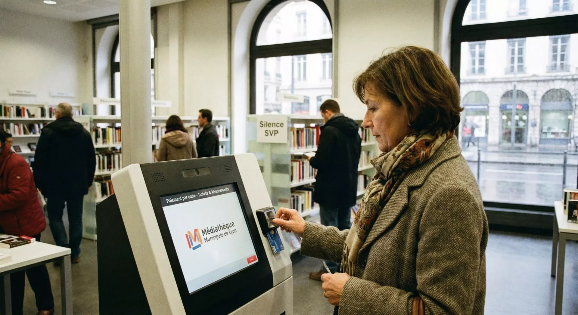 Femme utilisant une borne interactive dans une bibliothèque moderne avec des étagères et des fenêtres en arrière-plan.